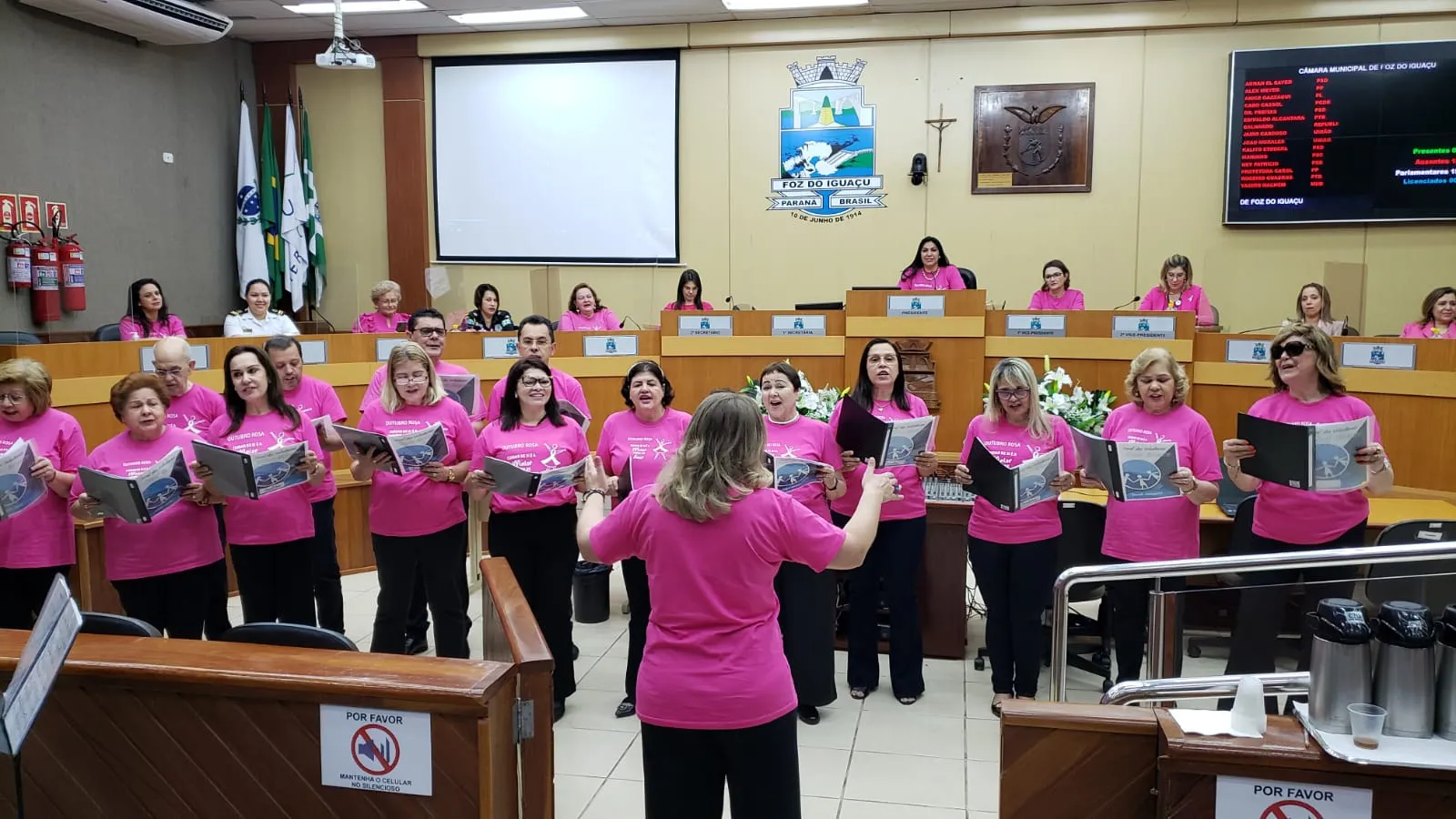 Coral Cisne Branco cantando de camiseta rosa na Câmara Municipal de Foz do Iguaçu, com a regente de costas em primeiro plano